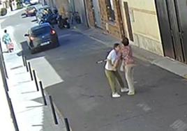 La asaltante, de blanco, forcejea con la víctima, que camina ayudada por una muleta. Al fondo, el coche del compinche.