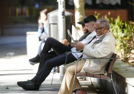 Un jubilado lee la prensa junto a dos jóvenes en el banco de un parque.