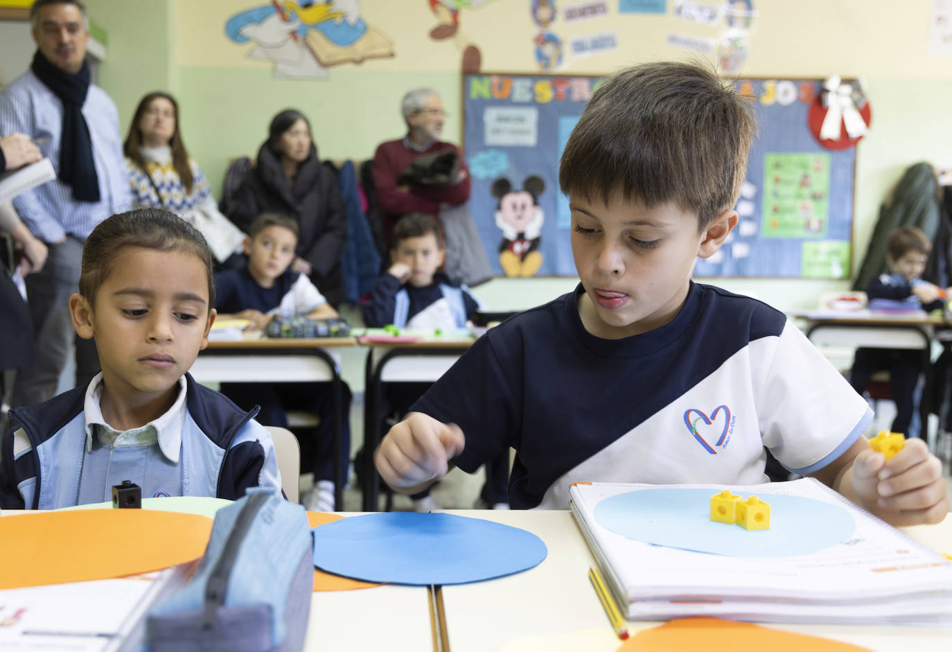 Dos alumnos de 2º de Primaria del centro Amor de Dios resuelven un problema matemático con objetos. Detrás, con un abrigo negro y un jersey granate, Josefa y Ramón observan a su nieto.