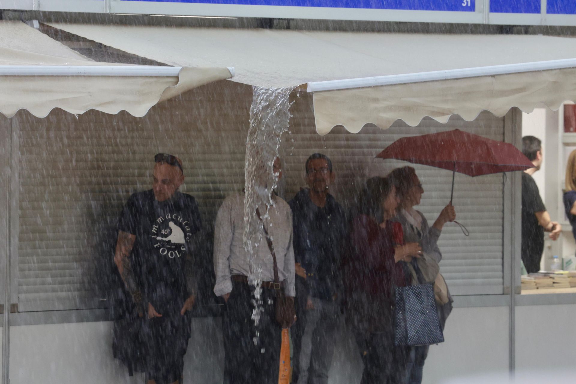 Algunos ciudadanos se refugian de la lluvia bajo los toldos de las casetas de la Feria del Libro instalada en la Plaza Mayor.