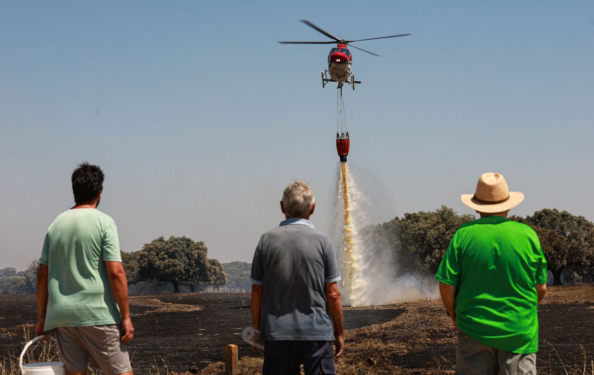Tres hombres observan cómo un helicóptero vierte agua para refrescar un terreno recién arrasado por un incendio forestal.