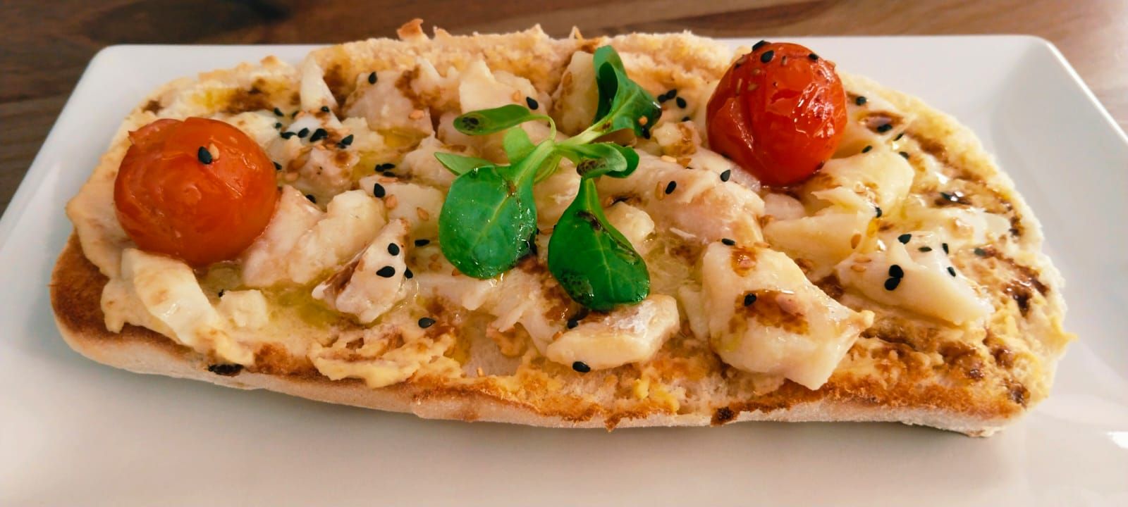 Tostada saludable en pan de chapata, integral o sin gluten. Bacalao portugués confitado a baja temperatura sobre hummus, acompañado de tomates cherry, canónigos y sésamo, aderezado con vinagreta de reducción casera de Oporto.