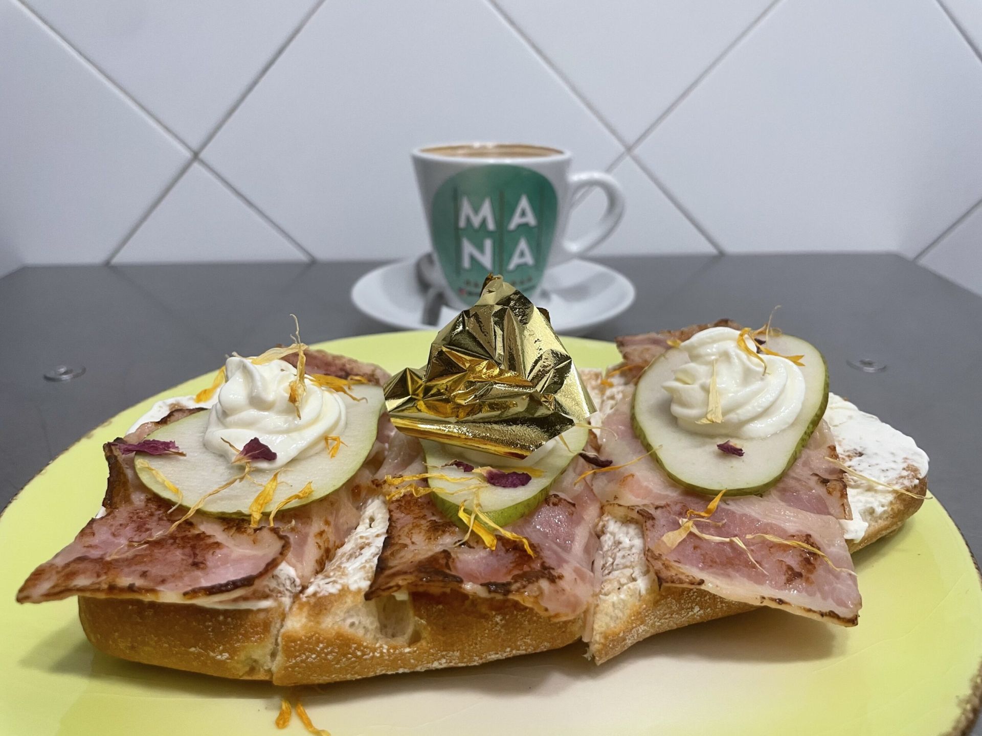 Una obra de arte que despierta los sentidos. Sobre base de pan de chapata, se extiende una suave mahonesa de ques. El bacon crunch añade el contraste perfecto. Finas láminas de pera para refrescan el paladar con su dulzor natural, mientras que un hilo sutil de miel de flores envuelve el conjunto con una fragancia floral que recuerda a un paseo entre jardines en primavera. Coronan esta creación, pétalos y papel de oro comestible.