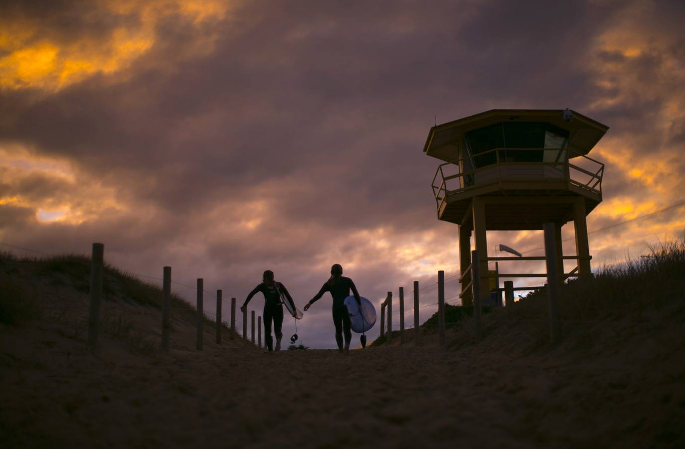 Un par de surfistas se dirigen a la cabeza de playa al lado de una cabaña de salvavidas al atardecer en Wanda Beach en Sydney.