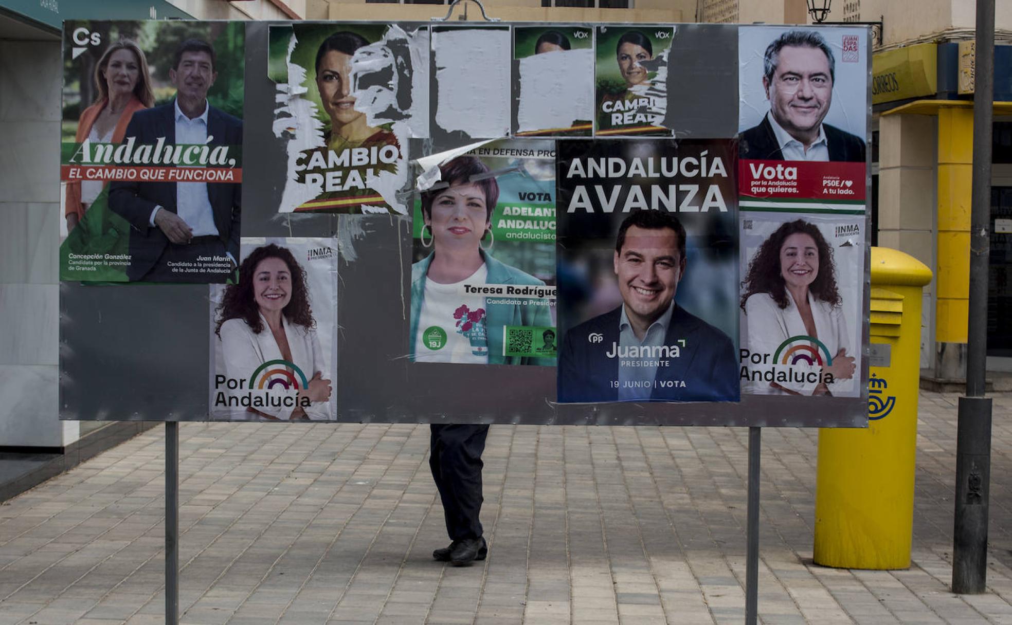 Una persona pasa tras un panel cubierto con carteles electorales de la campaña del 19J en la avenida Antonio Machado.