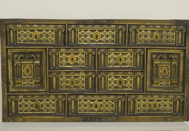 Escritorio de Salamanca en forma de caja rectangular, sin tapa frontal, con decoración arquitectónica de la muestra con placas de hueso, hueso torneado, policromado y dorado. La muestra con diez cajones y dos nichos cerrados por pequeñas puertas. No hay señal de que hubiera tenido una tapa frontal.