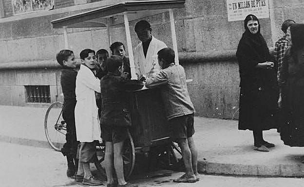 Años de Guerra Civil. Bajo la propaganda de las fuerzas sublevadas y el anuncio de la Lotería Pro Combatientes, un heladero atiende a unos niños junto al mercado en presencia de varias mujeres.
