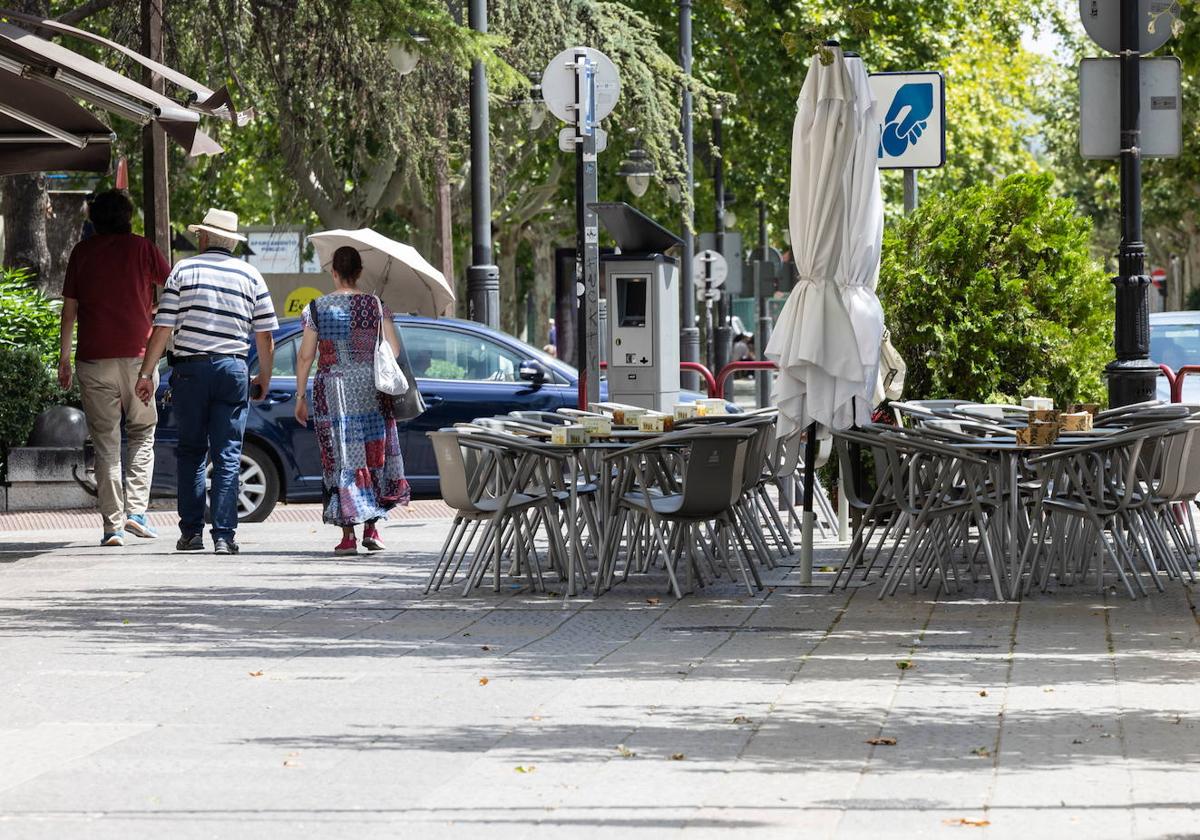 Dos personas caminan junto a una terraza vacía mientras se protegen del sol con un sombrero y un paraguas.