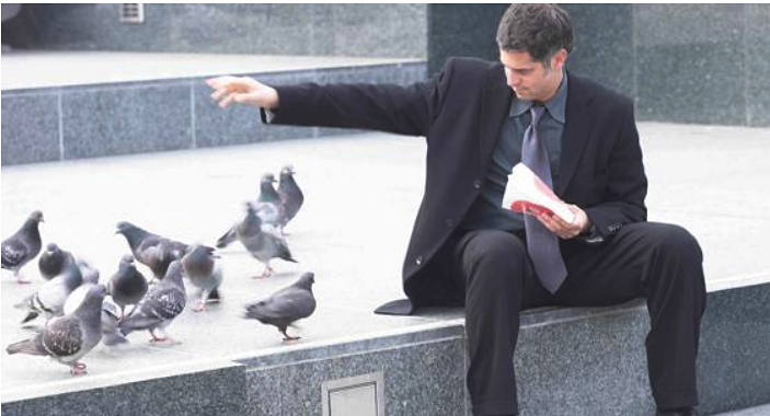 Un hombre sentado con las piernas abiertas da de comer a las palomas. 