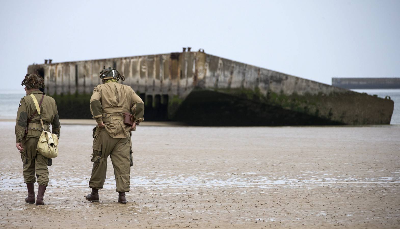 Varios entusiastas de la Segunda Guerra Mundial, con réplicas de uniformes estadounidenses de la época, caminan cerca de las ruinas del pontón de desembarco en la playa de Arromanches