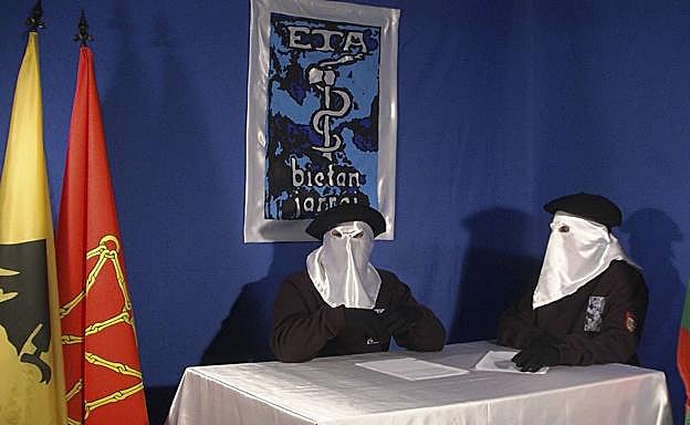 Etarras encapuchados sentados a una mesa debajo del logotipo de la banda terrorista. 