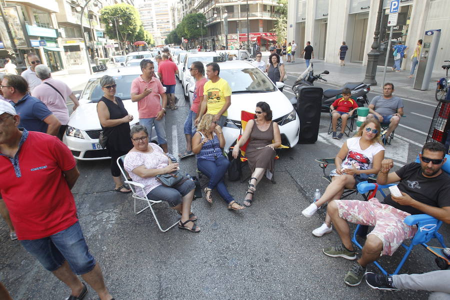 La huelga indefinida de taxistas en Valencia tiene como epicentro la calle Colón de Valencia. Allí, decenas de vehículo blancos están aparcados mientras los conductores tienen puesta toda la atención en la sede de Delegación del Gobierno. El único carril abierto es por el que circulan los autobuses de la EMT ante los taxistas que sentados en sillas plegables comentan, protestan y hace sonar el claxon. Unos conductores comparten café ante una mesa, mientras otros se muestran nerviosos con un cigarro en la mano. Ante la hora de comer, panes para los presenten y hasta se ha empezado un jamón entero. Las botellas de agua no han faltado ante las altas temperaturas que se registran en el centro de la ciudad.