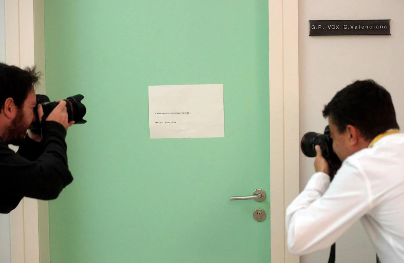 Dos fotógrafos toman una imagen de la puerta del Grupo Parlamentario de VOX en Les Corts Valencianes en cuya puerta se puede ver una cartel con el lema "Por prescripción facultativa, prohibido el paso hasta nueva orden". 