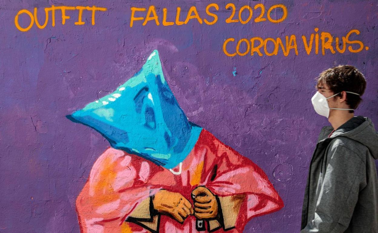 Una persona con mascarilla pasa frente a una pintura mural en referencia a las Fallas y el coronavirus. 