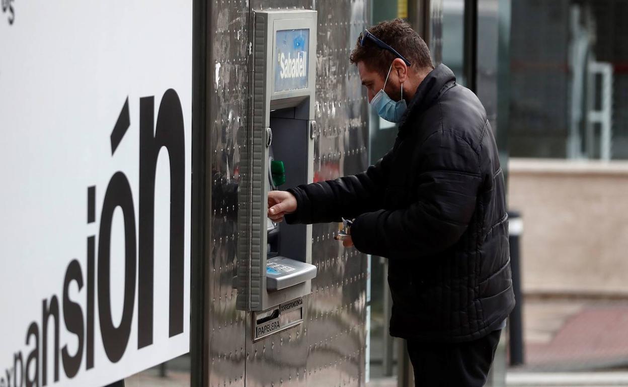 Un hombre saca dinero de un cajero automático, cubriendo la cara con una mascarilla.