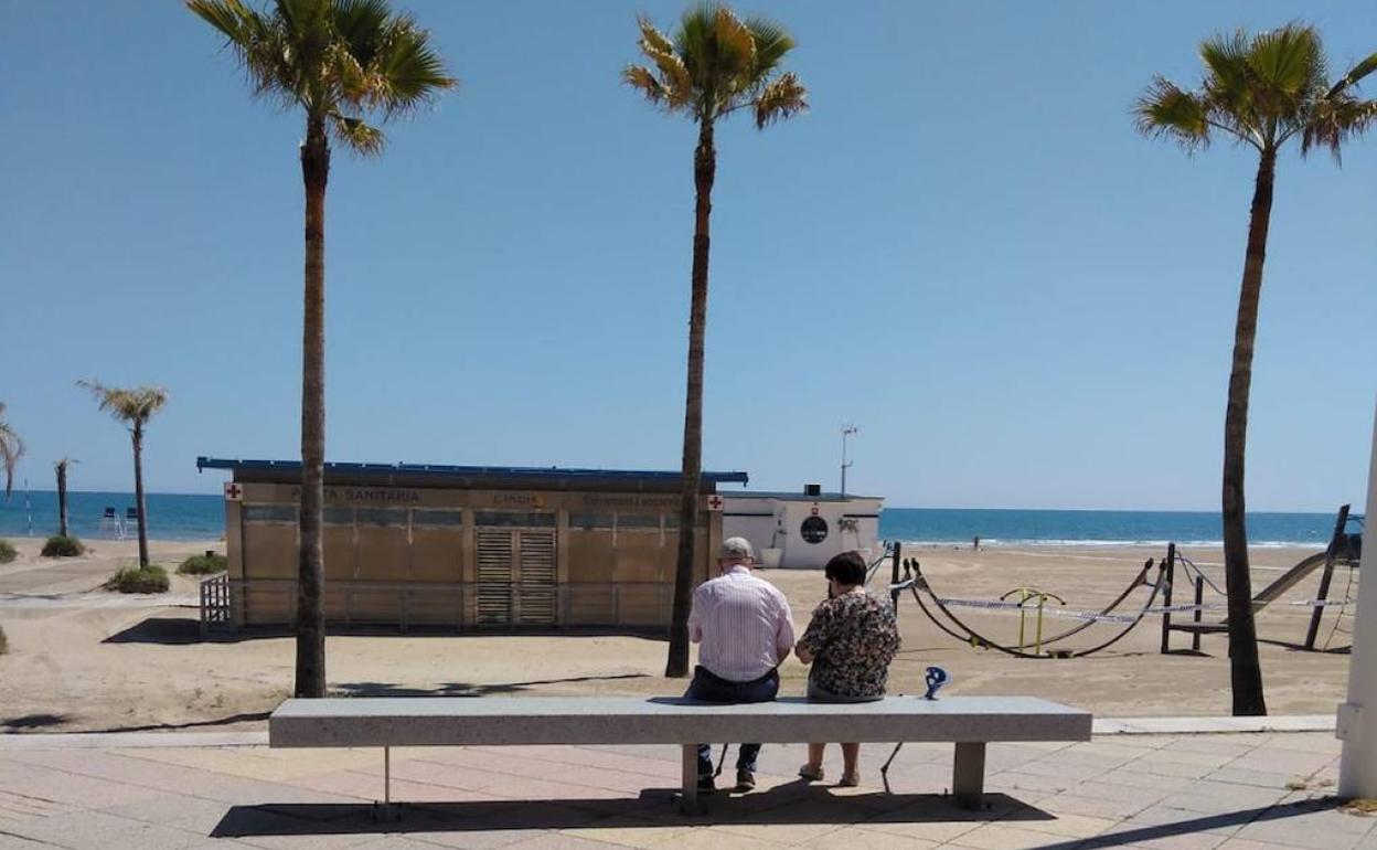 Una pareja sentada ayer por la mañana en un banco del paseo marítimo en la playa de Gandia frente a la posta sanitaria. 