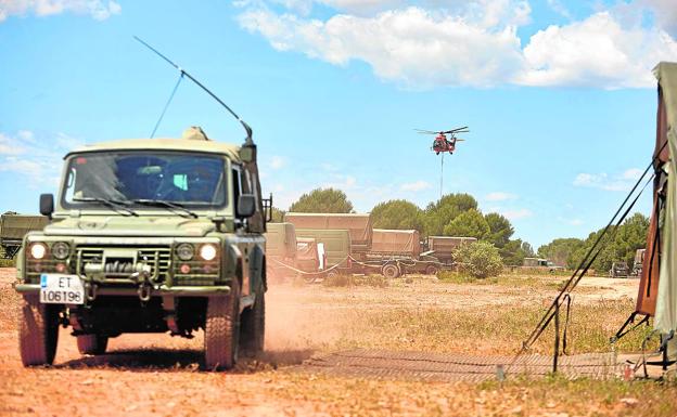 Imagen principal - Un vehículo con un helicóptero al fondo | Habitáculos de los soldados en el ejercicio | Dos miembros conversan con una soldado en el ejercicio.