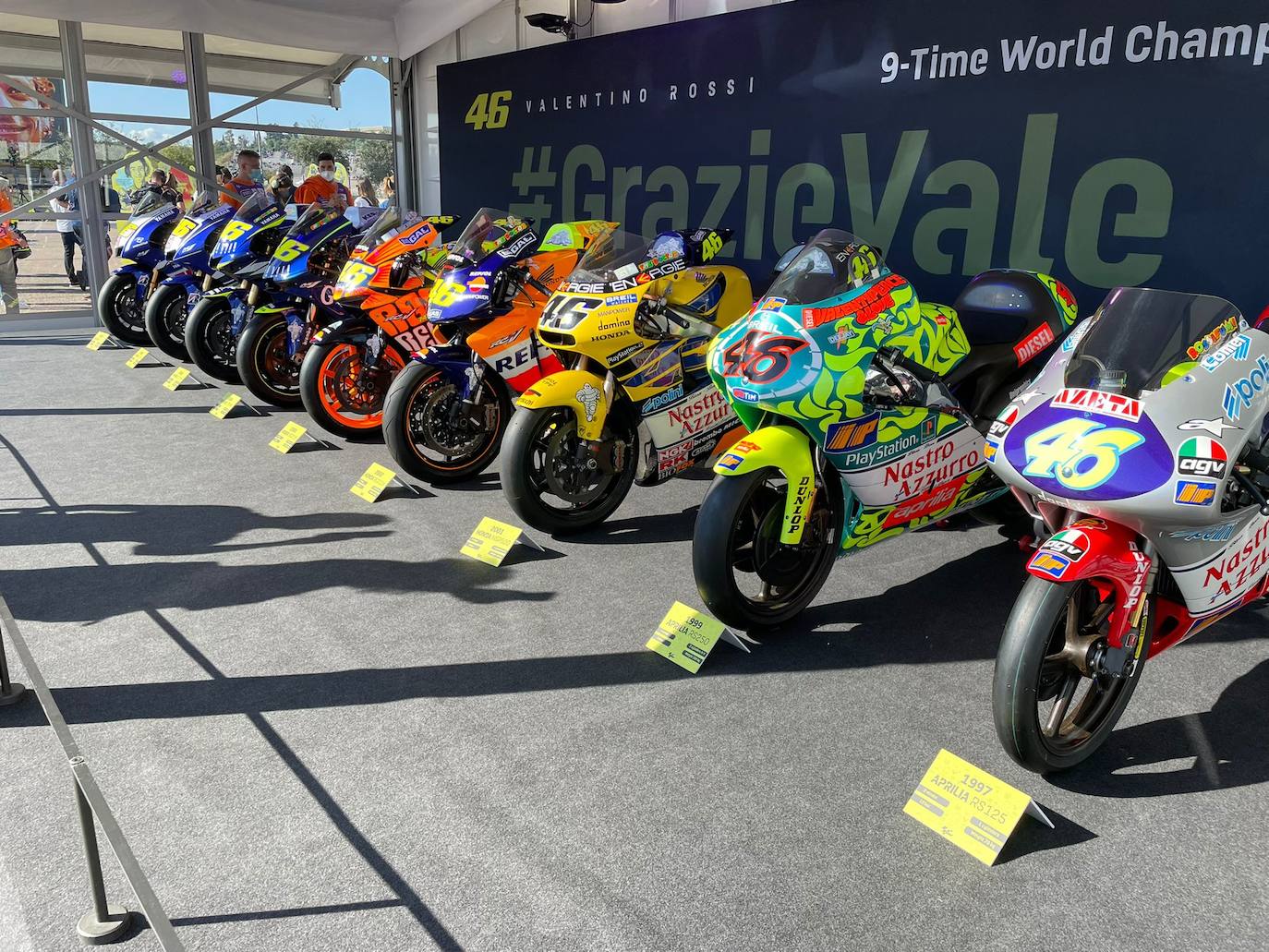 El paddock se ha teñido de amarillo este fin de semana. Como las gradas, que el sábado ya han expresado una muestra de intención de lo que será la carrera de este domingo. No es para nada exagerado afirmar que el 70% de los aficionados lucían merchandising de Valentino Rossi. Entre las muestras de homenaje que se le han dedicado por el momento, destaca la exposición de las nueve motos con las que ha sido campeón del mundo. Al lado está la 'Piazza Valentino Rossi', rotulada con una placa que simula ser de mármol y donde hay unos paneles amarilos que expresan los principales hitos del mito transalpino a lo largo de su carera. Más allá de esto, la mayoría de los camiones de los equipos tienen un panel, del mismo diseño, como muestra de gratitud al piloto por haber potenciado el Mundial de MotoGP durante sus más de 25 años de carrera. El propio piloto, en su mono, luce en la parte posterior este fin de semana un escueto pero representativo: 'Grazie'.