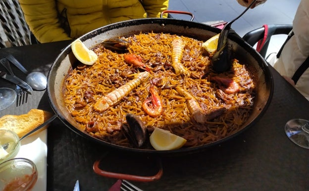 Imagen principal - Arriba, fideuà preparada en el Café Bar Diddley de Vilalonga. Abajo el plato típico de Gandia, elaborado en el Hogar del Pescador, en pleno puerto de Gandia. A la derecha los gerentes de Diddley, con una clienta. 