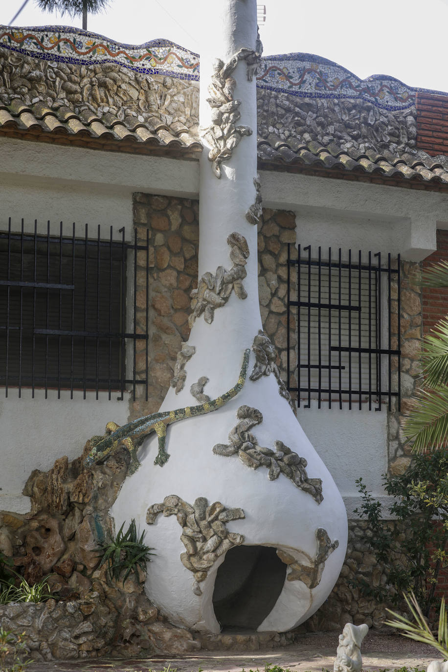 Una antigua chimenea ha sido transformada en ánfora gigante, con una apertura a modo de puerta concebida para albergar mascotas. Merodea un lagarto de cuya boca sale agua para regar las plantas.