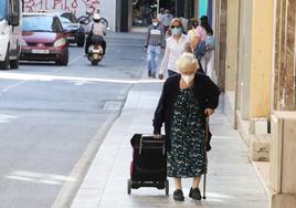 Una anciana camina con bastón y arrastrando un carro de la compra.