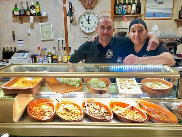 Imagen principal - Morell y Rosa Caudeli, junto al mostrador de tapas; un bocadillo de pisto con carne y una tapa de tortilla del día y habitas. 