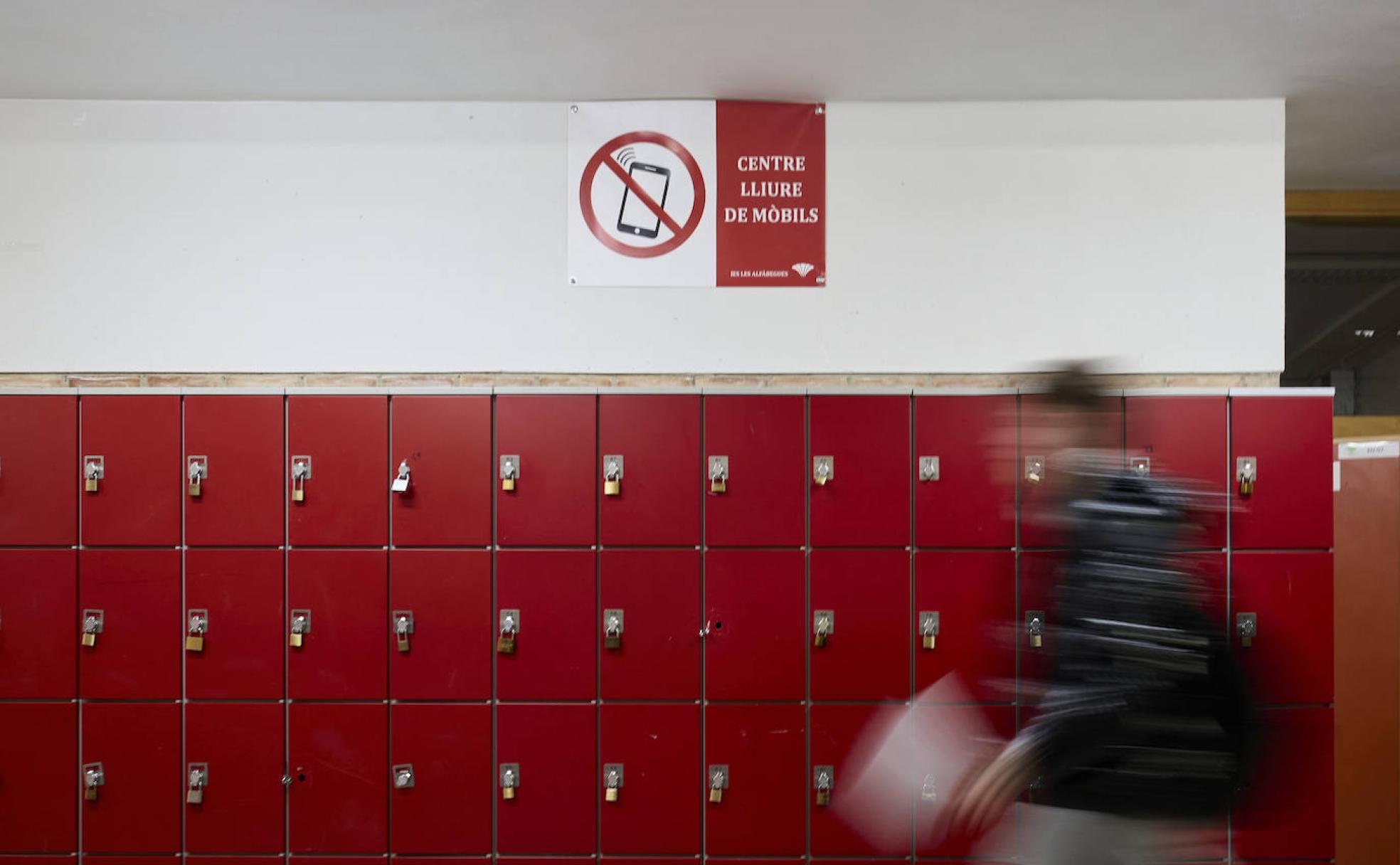 Un alumno atraviesa la zona de taquillas junto a uno de los carteles que limita el uso del teléfono móvil en el IES Les Alfàbegues de Bétera. 