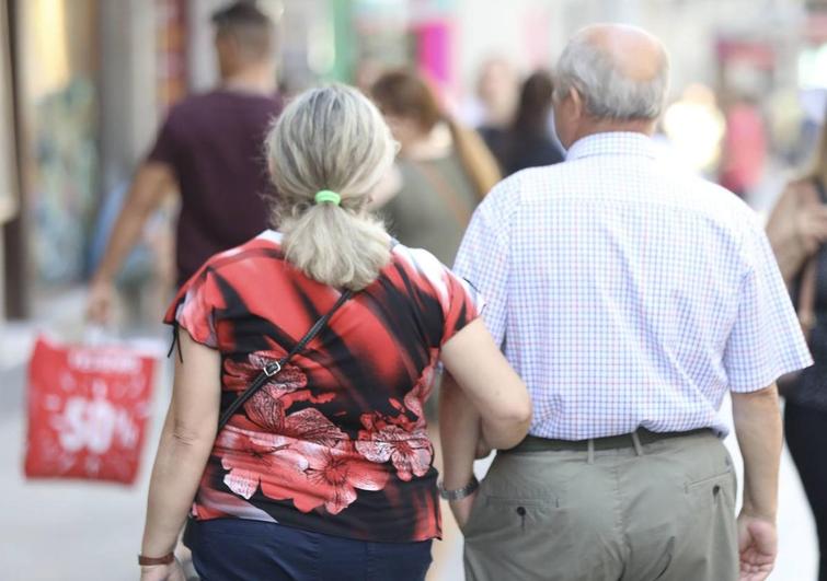 Una pareja de jubilados pasea por una calle comercial.