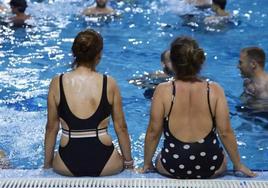 Dos mujeres sentadas al borde de una piscina, con otros bañistas.