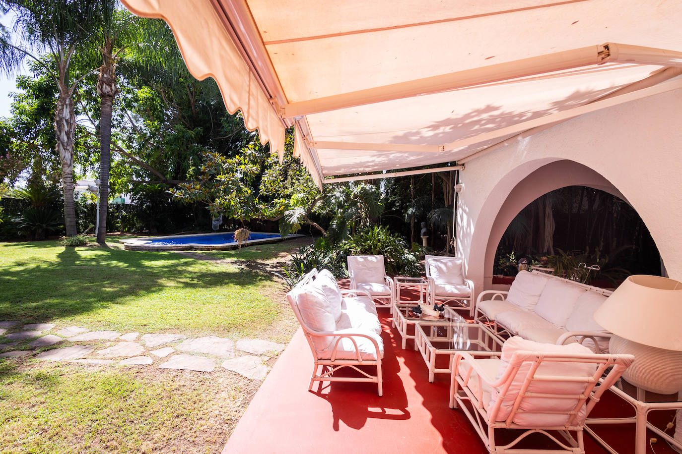 Zona de descanso en el jardín. Al fondo, la piscina, rodeada de vegetación.