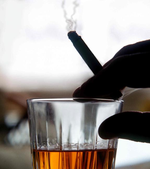 Un hombre toma un vaso de whisky mientras se fuma un cigarrillo.