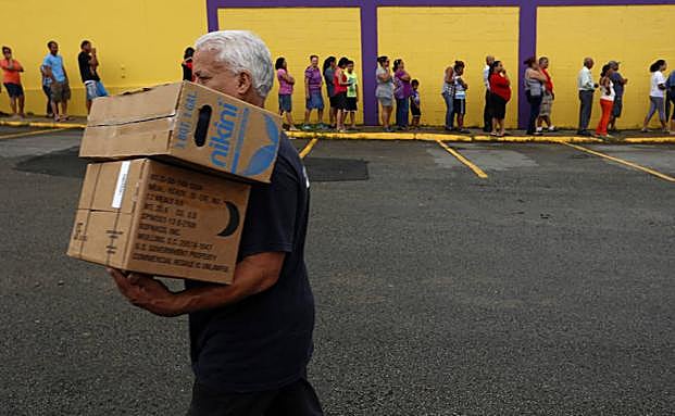 Un hombre camina con dos cajas de alimentos frente a una fila de personas damnificadas por el paso del huracán María. 