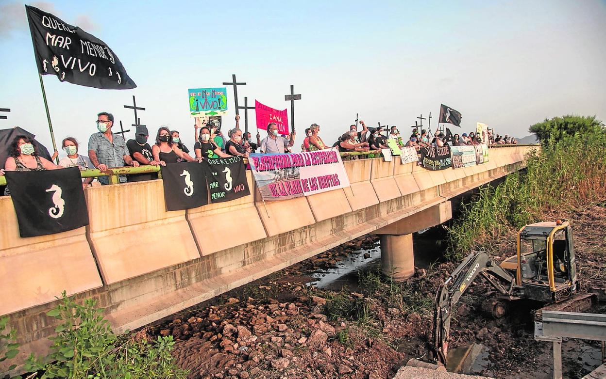 Protesta de vecinos en la rambla del Albujón, el mes pasado, para exigir la regeneración del Mar Menor. Debajo, una máquina retira cañas y sedimentos del cauce. 