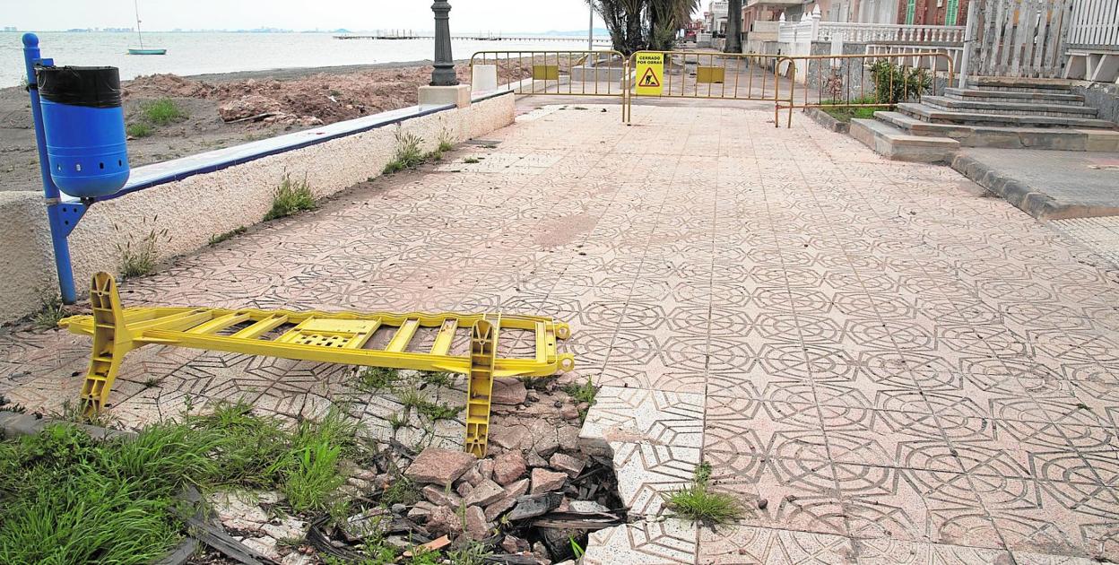 Boquete abierto y lleno de hierba, en el paseo marítimo de Los Urrutias y, al fondo, las obras que realiza el Estado para ampliar la zona de paso. 
