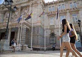 Dos chicas pasan frente al Palacio Consistorial mientras una pareja se besa con las banderas de fondo.