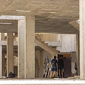 Dos hombres entran a un habitación construida bajo unas escaleras, mientras al fondo se ve otra chabola con ropa y varios muebles.