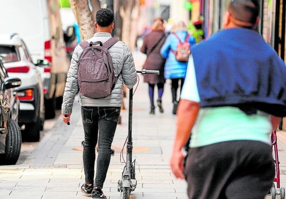 Un joven pasea por la calle Mayor de Alcantarilla mientras lleva un patinete con la mano.