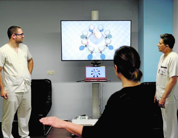 Una paciente realiza ejercicios con los brazos ayudada por una pantalla de realidad virtual, mientras el fisioterapeuta Juan Antonio López (derecha) y Alberto Villaescusa (alumno de Fisioterapia en prácticas) observan atentamente el desarrollo de la terapia. 