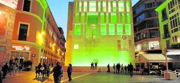 Varios grupos de personas pasean por la plaza de Belluga, anoche, con la fachada del Moneo iluminada en verde.