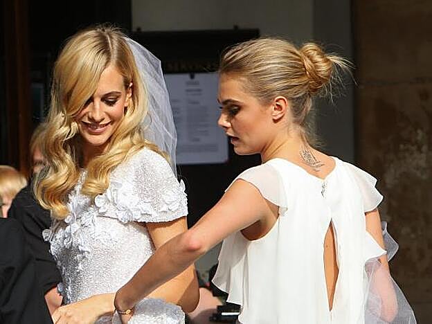 Poppy Delevingne con melena suelta ondulada y raya a un lado. De espaldas su hermana Cara Delevingne con moño de bailarina.