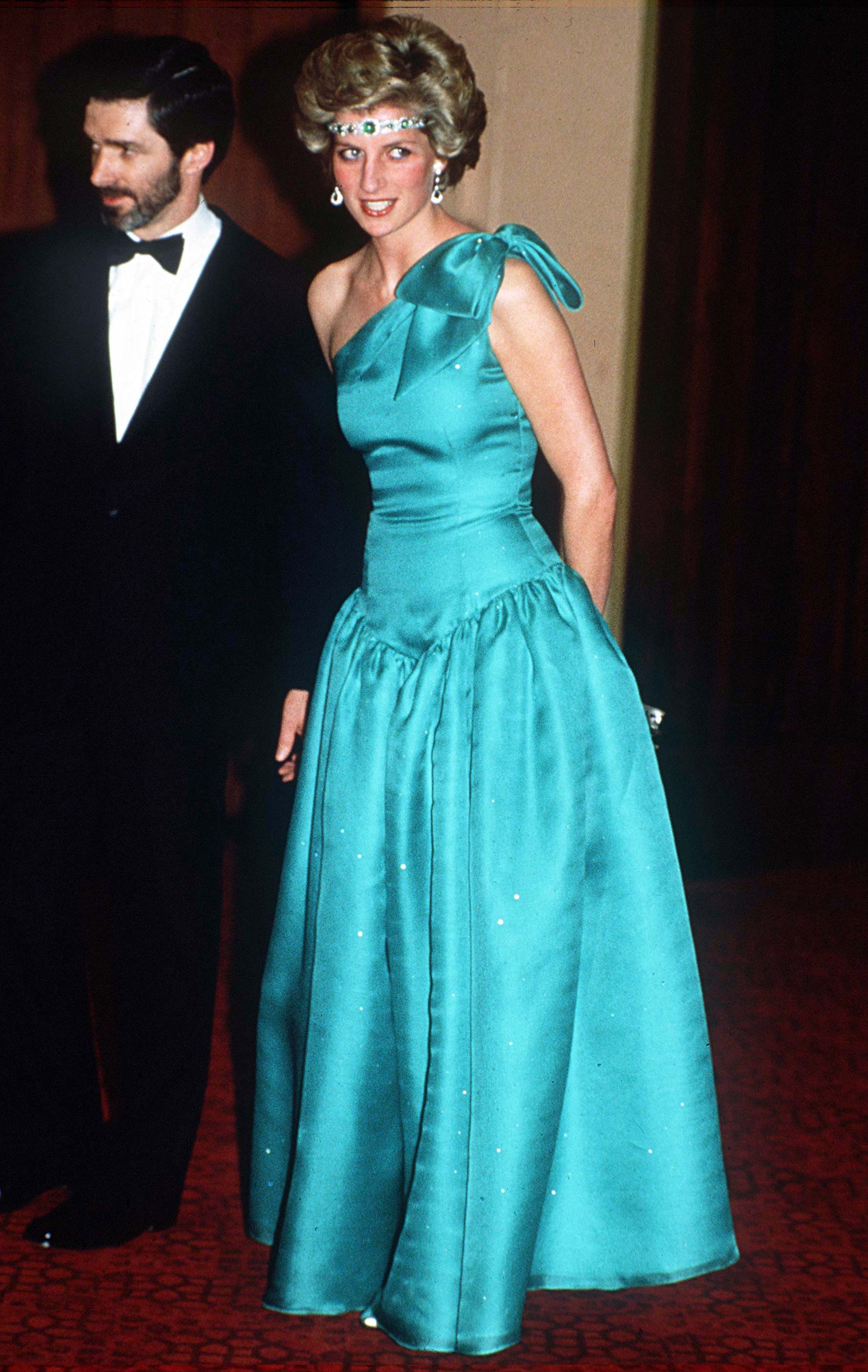 Diana de Gales con un vestido en color azul largo. Lleva de accesorios unos pendientes largos y una diadema en la frente.