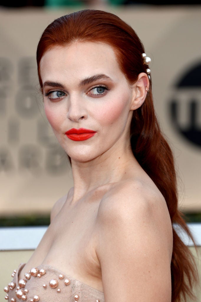 Madeline Brewer con el pelo suelto peinado hacia atrás y accesorio de perlas y unos labios rojos brillantes que contrastan con su piel blanca.