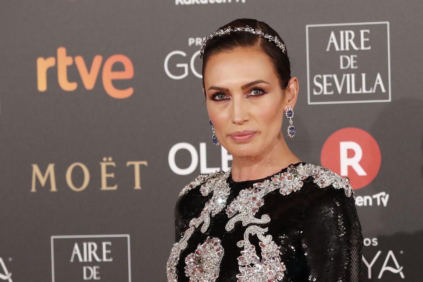 Nieves Álvarez con moño bajo pulido adornado con diadema joya, ojos enmarcados con eyeliner, sombras melocotón y azul glitter a juego con su vestido y pendientes y los labios nude.