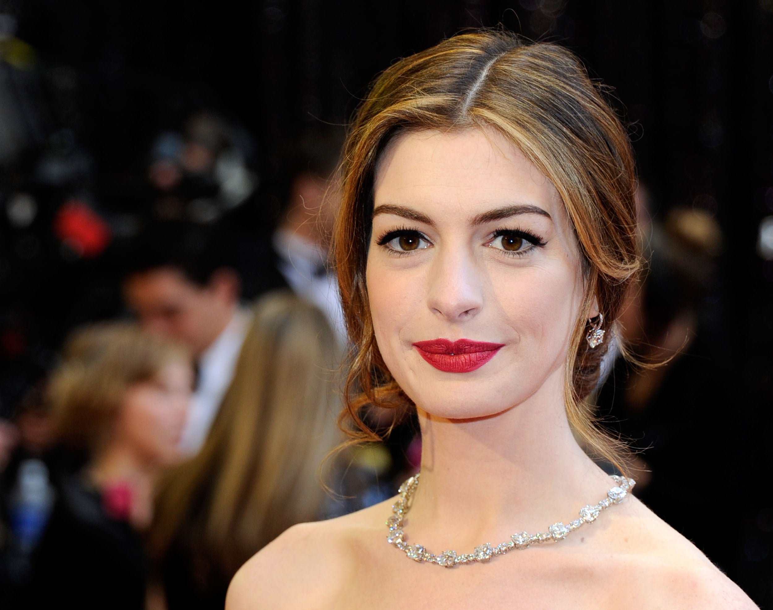 Anne Hathaway con recogido bajo ligeramente ladeado, mechones sueltos ondulados y raya en el medio en los Oscar 2011. Como maquillaje: labio s rojos y pestañas marcadas.