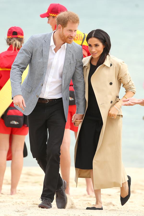 Meghan y Harry dieron un paseo por las playas de Melbourne para visitar a los socorristas, y allí vimos un nuevo cambio de look de la Duquesa de Sussex, que lució un sencillo pero acertadísimo vestido negro con detalle de botones dorados (en esta foto, no se aprecian) de la firma americana Club Monaco bajo su trench 'australiano' de Martin Grant, y cambió los tacones por unas bailarinas de Rothys (120 euros aproximadamente) mucho más apropiadas para caminar por la arena.