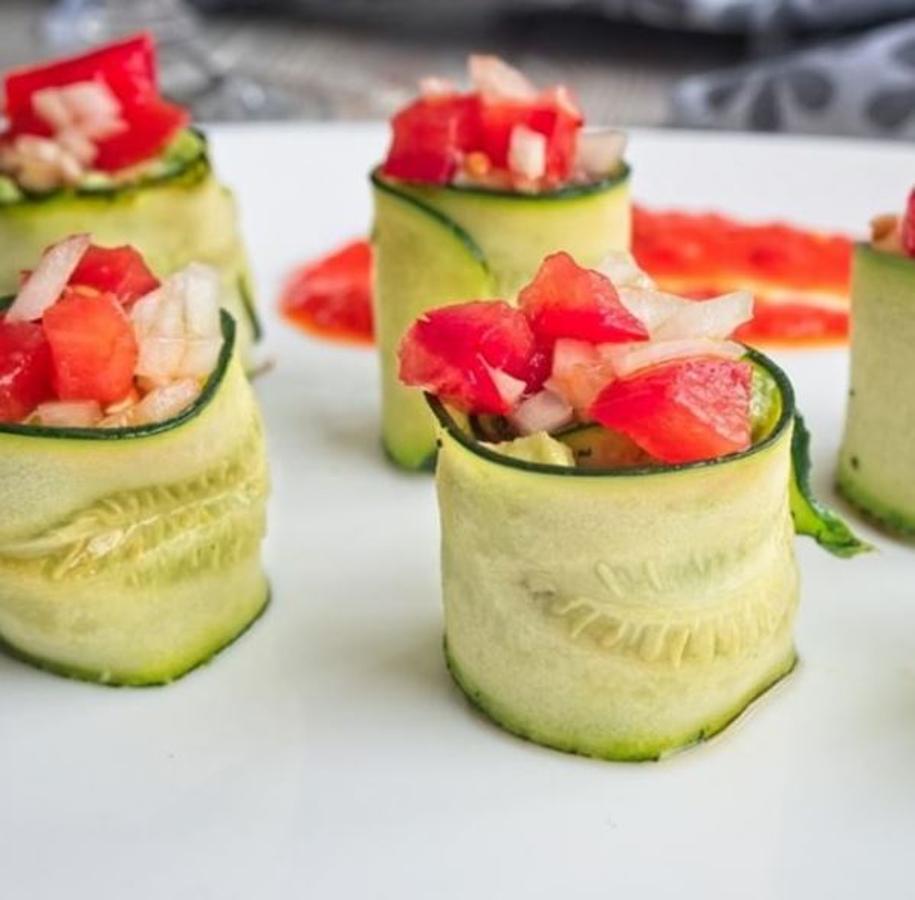 Ingredientes:-1 calabacín largo y estrecho-1 aguacate-1 tomate pequeño-1/4 cebolleta fresca-Paté de aceitunas negras-3 pimientos del piquillo-Limón-Sal-Pimienta negra-AOVEPreparación:Lava el calabacín, sécalo. Con ayuda de un pelador haz tiras, la primera deséchala, ya que solo será cáscara.Pon una plancha a calentar y unta ligeramente con aceite.Coloca las tiras de calabacín y deja que se hagan ligeramente. Solo un poco, ya que necesitamos que estén consistentes para enrollar. Es justo para quitarles la dureza del crudo.Pela el aguacate. Quita el hueso y aplasta con ayuda de un tenedor. Aliña con un poco de sal y pimienta negra. Además añade un poco de zumo de limón para que no se oxide.Untas las tiras de calabacín con el aguacate. Pon también un poco de paté de aceitunas.Enrolla con cuidado y pon hacia arriba el rollito.Corta la cebolleta y el tomate en cuadraditos pequeños y aliña con un poco de sal y aceite.Coloca esta mezcla sobre los rollitos con cuidado.Pon los pimientos en el vaso de la batidora y tritura junto con un par de cucharadas de aceite, un poco de sal y un poco de pimienta negra molida. Tritura hasta dejar una crema.Sirve los rollitos con un poco de crema de pimientos al lado y así podrás untar los rollitos si te apetece.