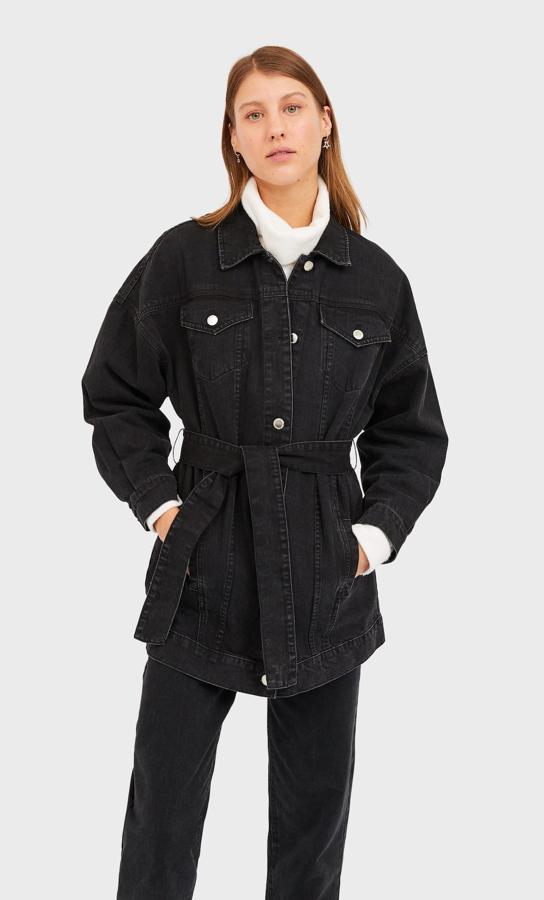 Chaqueta vaquera negra larga con cinturón de Stradivarius. Costaba 35,99 euros y está rebajada a 29,99 euros.