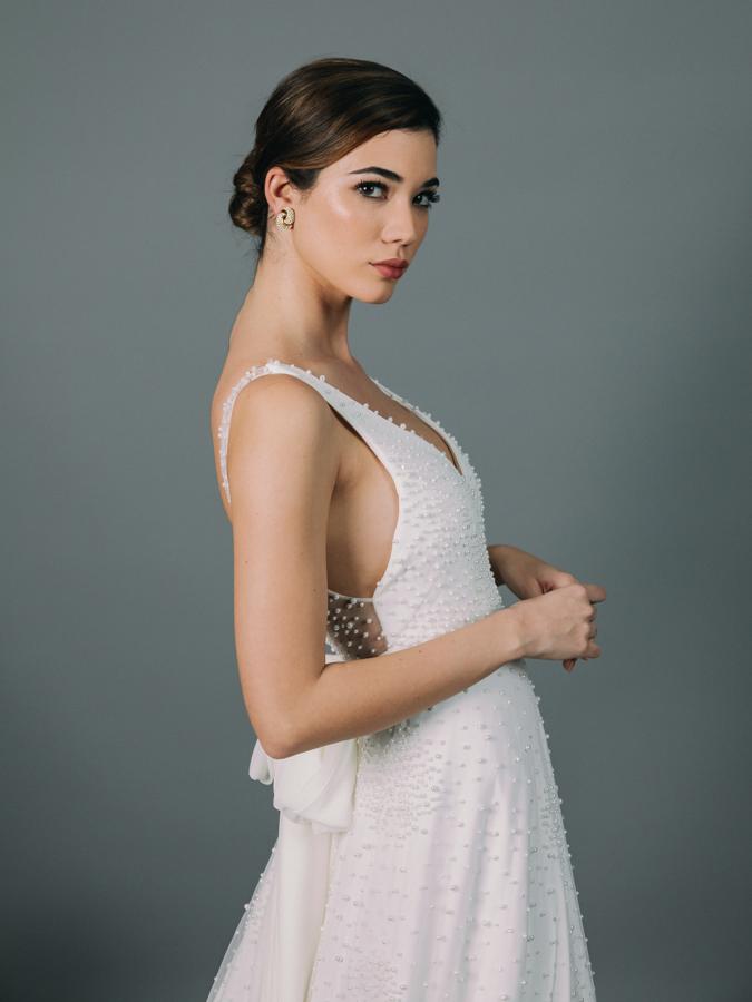 Vestido de novia con escote en pico y cuerpo entallado. Corte en la cintura y falda de capa con cola. Confeccionado en tul bordado en perlas y cristal. Detalle de lazada en la espalda en gasa natural.