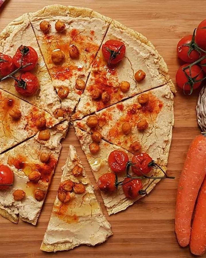 Ingredientes para la base:150 gr de harina de maíz1 pizca de salPimienta negraAgua tibiaIngredientes para el hummus:2 botes de garbanzos cocidos1 cucharada de tahini1 pizca de salZumo de medio limón1 cucharadita de comino en polvo1 diente de ajo1cucharada de AOVEIngredientes para los garbanzos especiados:GarbanzosCúrcumaPImentón dulcePimentón picante1 pizca de salPreparación:- Para hacer la base, en un bol añadimos la harina de maíz, la sal y la pimienta al gusto. Mezclamos y vamos añadiendo agua tibia hasta formar una masa compacta y manejable.- En una sartén antiadherente echamos un poquito de AOVE e impregnamos bien toda la base con ayuda de papel de cocina.- Colocamos la bola de la masa y la vamos extendiendo con ayuda de una espátula desde el interior hacia los bordes de la sartén. Debe quedar muy fina. Si sobra masa, se retira.- Una vez extendida, colocamos una tapa a la sartén y cocinamos a fuego medio.- Para el hummus, en un procesador de alimentos echamos todos los ingredientes y trituramos. Añadimos agua si fuera necesario.- Para los garbanzos especiados, los impregnamos con las especias al gusto y los colocamos en una bandeja de horno. Horneamos a 180ºC durante 40 minutos.- Extendemos el hummus sobre la base de la pizza y ponemos por encima los garbanzos especiados. Podemos añadirle unos tomatitos previamente horneados, pimentón y AOVE.