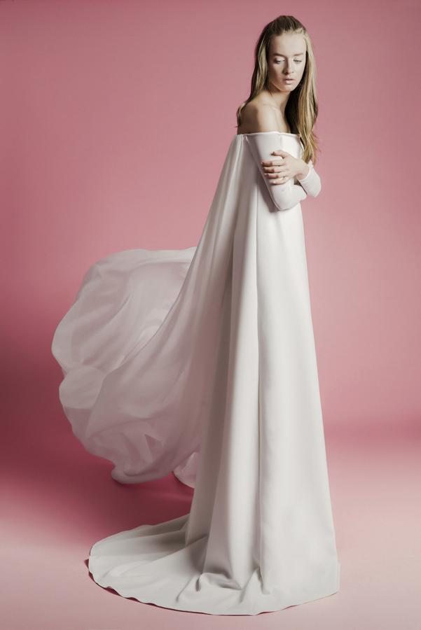 Vestido de novia recto con cola y capa, con escote barco y manga larga ajustada.